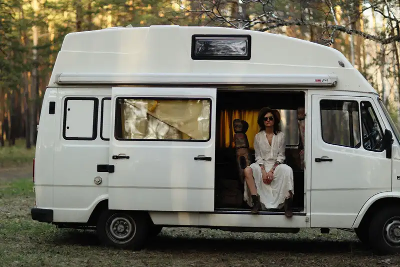 A woman wearing a white dress sits in a camper van parked at a forest campsite.